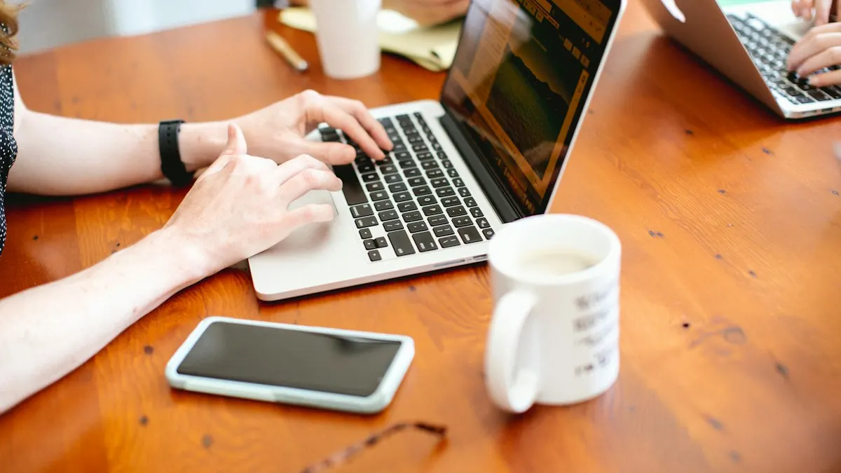 Hände tippen auf der Tastatur eines Laptops auf einem Holztisch, daneben liegen ein Mobiltelefon und ein Becher.