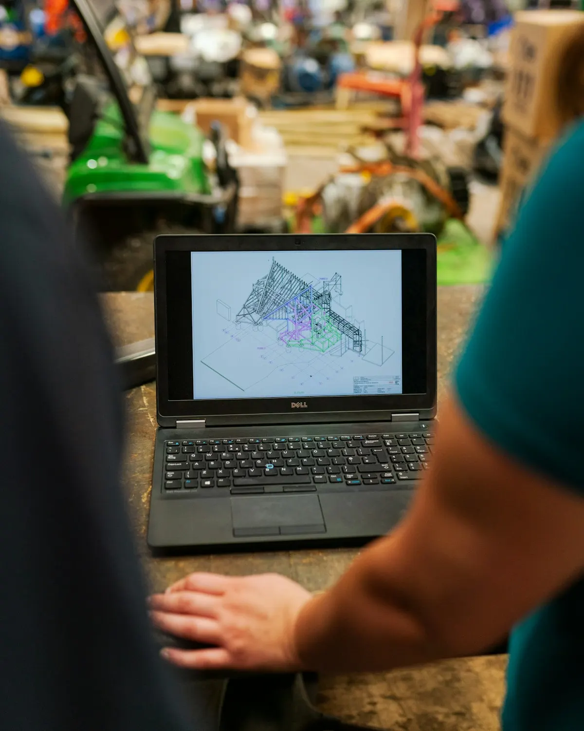 Laptop mit einer technischen Konstruktionszeichnung auf einer Werkbank in einer Werkhalle, davor ruht eine Hand auf der Arbeitsplatte.