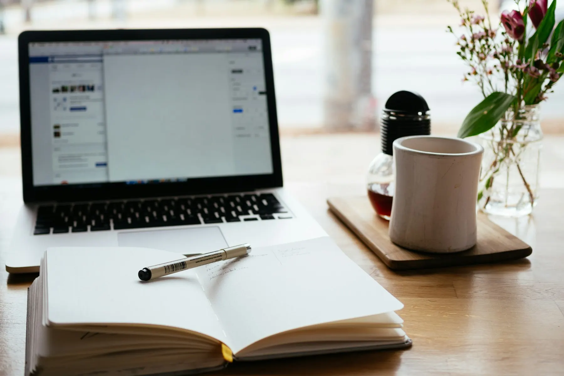 Aufgeschlagenes Notizbuch mit Stift vor einem Laptop, daneben ein Becher und eine Vase mit Blumen auf einem Holztisch.